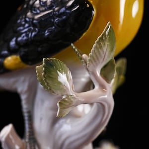 May include: A detailed, close-up of a porcelain bird figurine. The bird has black and yellow plumage, perched on a white branch with green and white leaves. The figurine is set against a black background.