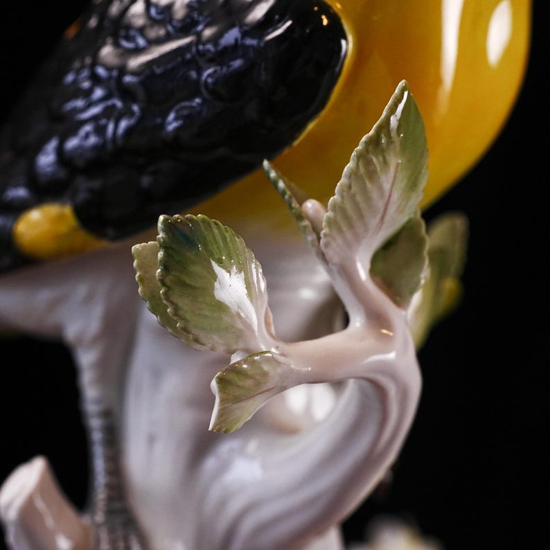 May include: A detailed, close-up of a porcelain bird figurine. The bird has black and yellow plumage, perched on a white branch with green and white leaves. The figurine is set against a black background.