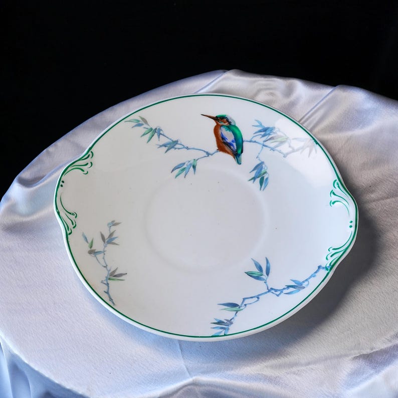 May include: White ceramic plate with a green rim and decorative handles. The plate features a colorful illustration of a bird perched on a branch with blue leaves. The bird has orange, green, and blue feathers.