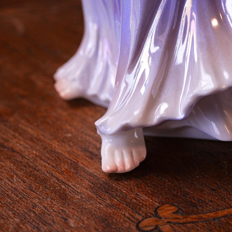 May include: A close-up of a porcelain figurine's feet and flowing gown. The gown transitions from a soft lavender to white, with subtle iridescent highlights. The figurine's feet are delicately painted with pink tones.
