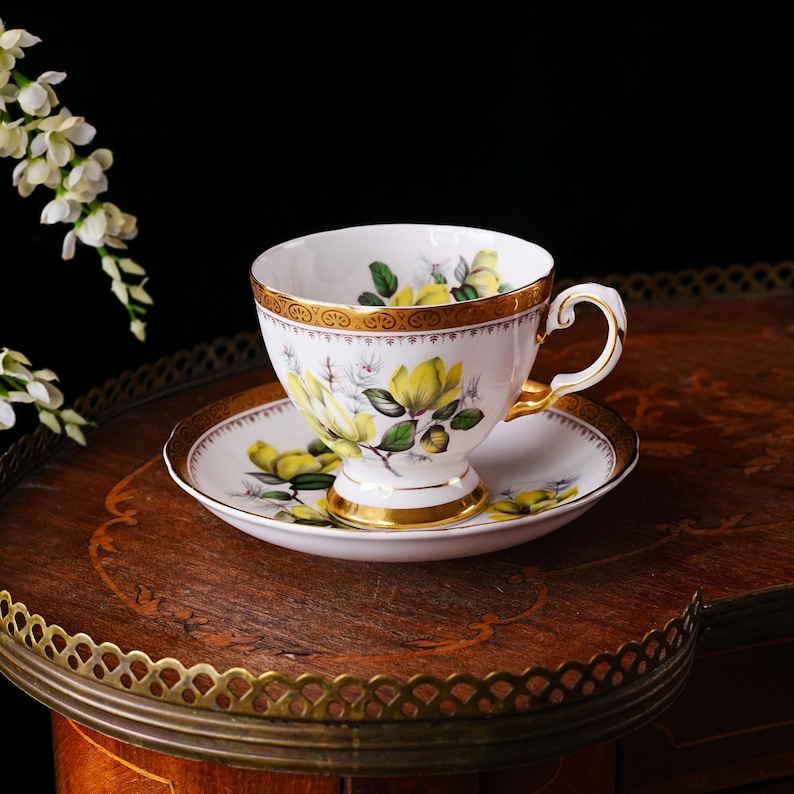 Peut inclure: Une tasse et une soucoupe blanches avec un motif floral jaune et des bordures dor&eacute;es. La tasse a une anse incurv&eacute;e et repose sur une soucoupe assortie. L'ensemble est pos&eacute; sur une table en bois.