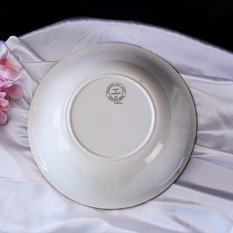 May include: A white ceramic plate with a cream-coloured rim. The underside has a circular stamp with text: "Ridgways Old Ivory, Made in England, Bedford Ware". The plate is on a white satin background.