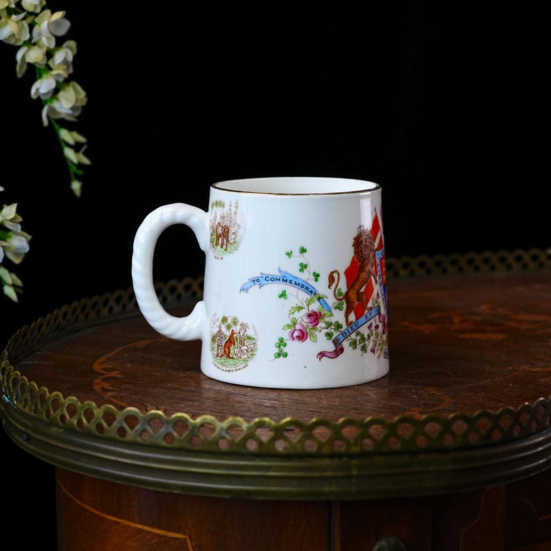 Op de afbeelding: Witte herdenkingsmok met een gouden rand en een decoratief handvat. De mok heeft kleurrijke illustraties van een leeuw, een vlag en bloemmotieven, met de tekst "TO COMMEMORATE". De mok staat op een houten tafel.