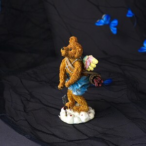 Puede incluir: Una figura de oso de peluche marr&oacute;n al estilo de Cupido, con alas multicolores, un pa&ntilde;o azul y un carcaj con flechas en la espalda. El oso se apoya sobre una base de nube blanca con una peque&ntilde;a flecha colocada verticalmente frente a sus pies.