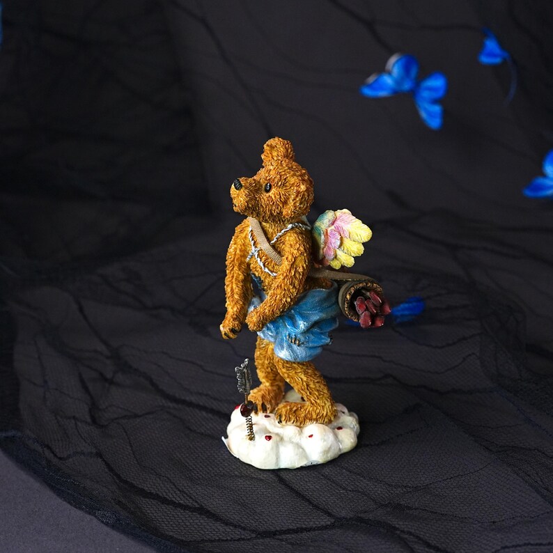 Puede incluir: Una figura de oso de peluche marr&oacute;n al estilo de Cupido, con alas multicolores, un pa&ntilde;o azul y un carcaj con flechas en la espalda. El oso se apoya sobre una base de nube blanca con una peque&ntilde;a flecha colocada verticalmente frente a sus pies.