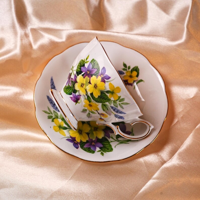 K&ouml;nnte beinhalten: Ein wei&szlig;es Tassen- und Untertassen-Set mit einem Blumendesign mit gelben und lila Blumen. Die Tasse und die Untertasse haben einen goldenen Rand. Das Set steht auf einer pfirsichfarbenen Satin-Oberfl&auml;che.