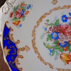 Puede incluir: Un plato de cer&aacute;mica blanca con un ramo floral central de flores rojas, azules y amarillas rodeado por un borde de filigrana dorada. El plato incluye una secci&oacute;n de color azul cobalto vibrante con volutas doradas y un peque&ntilde;o grupo floral.