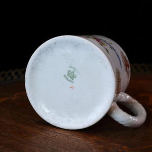 Op de afbeelding: Witte porseleinen mok met een gedraaide handgreep, versierd met bloemen- en architecturale patronen in rood, blauw en groen. De onderkant van de mok toont de woorden "AYNSLEY ENGLAND" en een klein embleem.