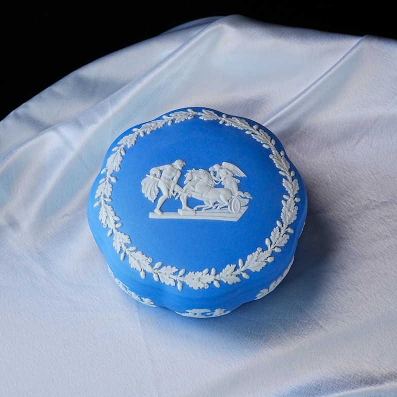 May include: A round blue ceramic trinket box featuring white relief ornamentation. The lid displays a central scene of a figure with a horse and a winged figure, all encircled by a detailed white oak leaf and acorn border pattern.