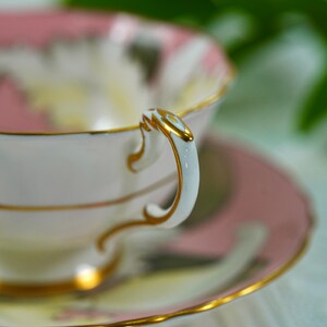 May include: A close-up of a vintage teacup and saucer set. The teacup and saucer are white with pink floral designs and gold trim. The handle of the teacup is white with gold accents. The saucer is pink with a gold rim.