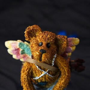 Puede incluir: Una figura de oso de peluche con textura marr&oacute;n y alas multicolores con detalles en amarillo, rosa y verde. El oso tiene ojos negros, nariz negra y est&aacute; adornado con una correa color canela y un cord&oacute;n plateado cruzado sobre el pecho.