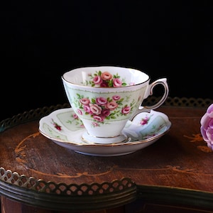 K&ouml;nnte beinhalten: Ein Vintage-Tee-Set, bestehend aus einer Tasse und einer Untertasse. Die wei&szlig;e Porzellantasse und Untertasse zeigen ein Blumenmuster aus rosa Rosen und gr&uuml;nem Laub. Das Set hat einen Goldrand und steht auf einer Holzoberfl&auml;che.