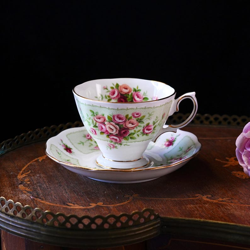 K&ouml;nnte beinhalten: Ein Vintage-Tee-Set, bestehend aus einer Tasse und einer Untertasse. Die wei&szlig;e Porzellantasse und Untertasse zeigen ein Blumenmuster aus rosa Rosen und gr&uuml;nem Laub. Das Set hat einen Goldrand und steht auf einer Holzoberfl&auml;che.