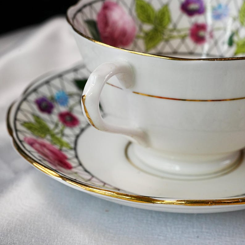 Op de afbeelding: Een witte theekop en schotel set met gouden rand. De handgreep en rand van de kop zijn goudkleurig. De schotel en de binnenkant van de kop hebben een bloemenpatroon met roze, paarse en blauwe bloemen. De set staat op een wit oppervlak.