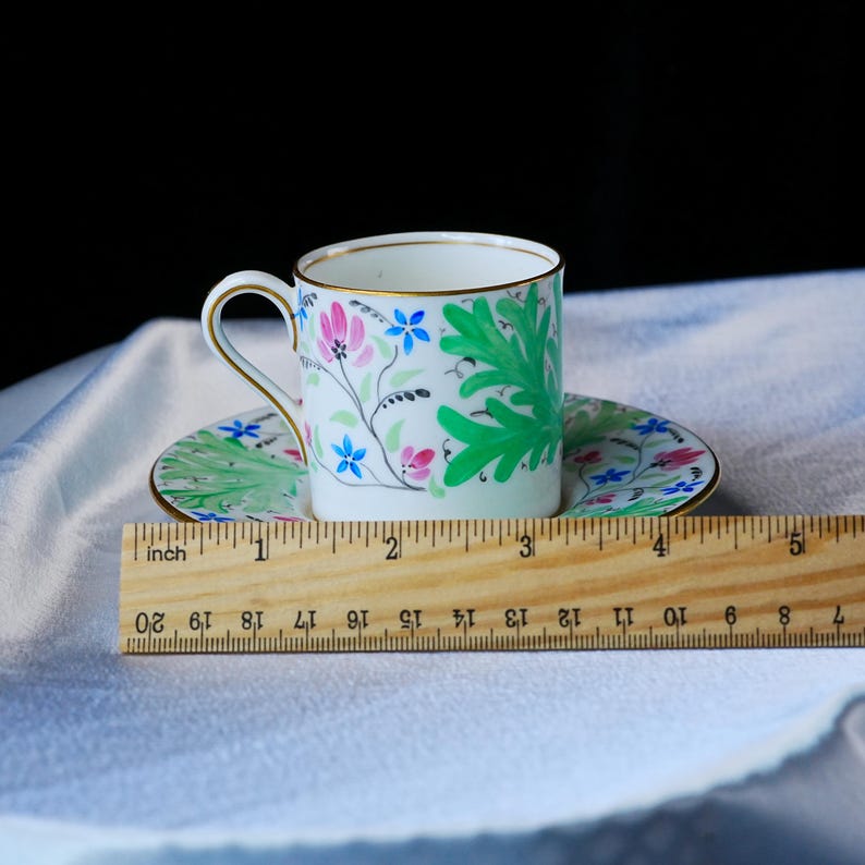May include: A white porcelain teacup and saucer set featuring hand-painted green leaves, pink flowers, and blue blossoms with gold trim. A wooden ruler placed in front shows the saucer has a diameter of approximately 5.5 inches.