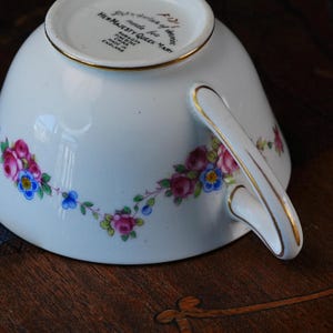 Op de afbeelding: Een witte theekop van fijn beenderporselein met een bloemenslinger van roze rozen en blauwe vergeet-mij-nietjes, afgewerkt met een gouden rand op het oor en de bovenkant. De onderkant bevat de tekst: By Appointment of Service, Made for Her Majesty Queen Mary, Paragon, Fine Bone China, Made in England.