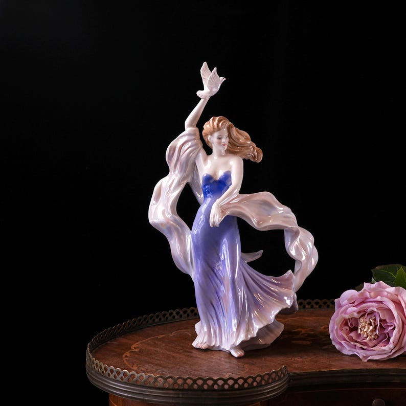 May include: A porcelain figurine of a woman in a flowing lavender gown with a blue bodice, holding a white dove. Her long, flowing hair is a light brown colour, and she is draped in a white scarf. The figurine is on a wooden table with a rose.