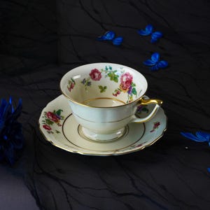 Op de afbeelding: Een crèmekleurige porseleinen theekop en schotelset met een delicaat bloemmotief van roze rozen en groene bladeren. De rand van de kop en de schotel is geaccentueerd met een dunne gouden bies en de kop is voorzien van een goudkleurig oortje.