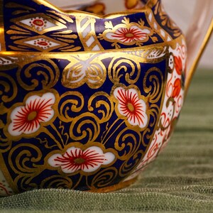 May include: Close-up of a decorative ceramic pitcher with a handle. The pitcher features a vibrant design with a navy blue background, gold accents, and floral patterns in red and white. The rim has a scalloped edge.