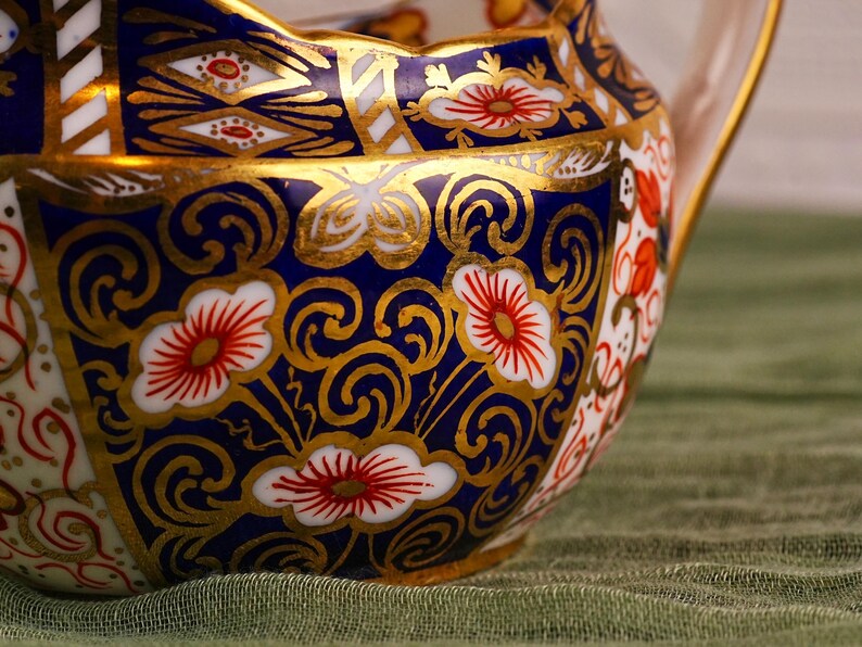 May include: Close-up of a decorative ceramic pitcher with a handle. The pitcher features a vibrant design with a navy blue background, gold accents, and floral patterns in red and white. The rim has a scalloped edge.