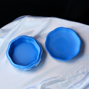May include: A bright blue ceramic trinket box with a scalloped edge and a matching lid. The exterior of the base features white relief detailing. The interior of both the base and the lid is a solid, matte blue finish, showing a circular shape.