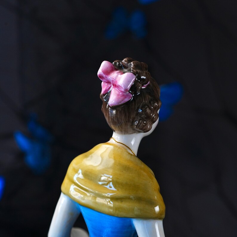 May include: A porcelain figurine of a figure with brown hair styled in an updo, adorned with a glossy pink bow. The figure wears a mustard yellow shawl draped over the shoulders and a bright blue garment, featuring a thin gold-toned chain necklace.
