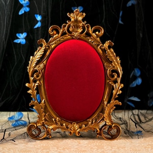 Puede incluir: Marco de fotos ovalado dorado con interior de terciopelo rojo. El marco tiene intrincados adornos y una parte superior decorativa. El marco es independiente, adecuado para exhibir fotos o obras de arte. El fondo presenta acentos de mariposas azules.