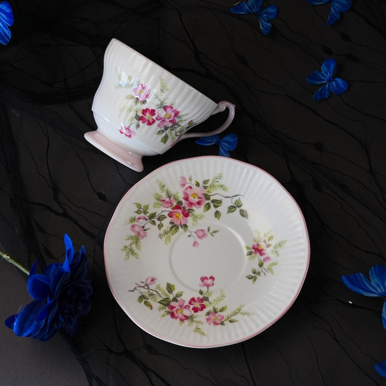 May include: A white porcelain teacup and matching saucer featuring a ribbed texture and a delicate floral pattern of pink roses with green leaves. The teacup has a light pink handle and base, while the saucer edge is trimmed in soft pink.