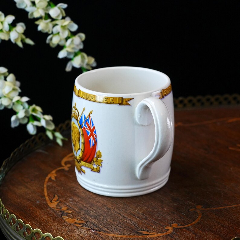 May include: White ceramic mug with a curved handle. The mug features a design with a crown, flags, and the word "JUBILEE" in gold lettering. The mug is sitting on a wooden surface.