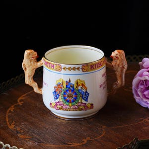 Puede incluir: Una taza de cer&aacute;mica blanca con dos asas esculpidas. La taza presenta un borde dorado y un dise&ntilde;o de escudo de armas colorido con banderas y texto. Las asas est&aacute;n esculpidas para parecer figuras.