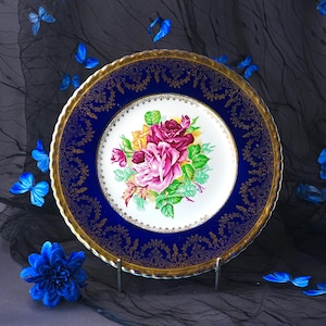 May include: A round ceramic plate featuring a central white medallion with a bouquet of pink, yellow, and deep red roses with green leaves. The wide border is deep cobalt blue with intricate gold filigree patterns and a scalloped gold rim.