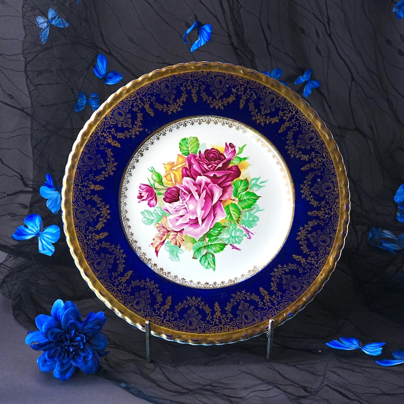May include: A round ceramic plate featuring a central white medallion with a bouquet of pink, yellow, and deep red roses with green leaves. The wide border is deep cobalt blue with intricate gold filigree patterns and a scalloped gold rim.