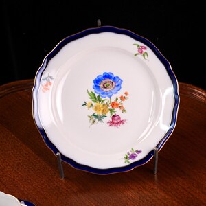 Puede incluir: Plato de porcelana blanca con borde ondulado, borde azul y ribete dorado. El plato presenta un dise&ntilde;o floral central con una gran flor azul y flores m&aacute;s peque&ntilde;as amarillas, naranjas y rosas. El plato se exhibe sobre un soporte met&aacute;lico.