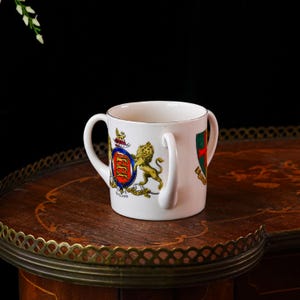 Puede incluir: Taza de cerámica blanca con dos asas, con un escudo heráldico con un león y un escudo. La taza tiene un borde dorado y está colocada sobre una mesa de madera oscura. El escudo incluye la palabra "ENGLAND".
