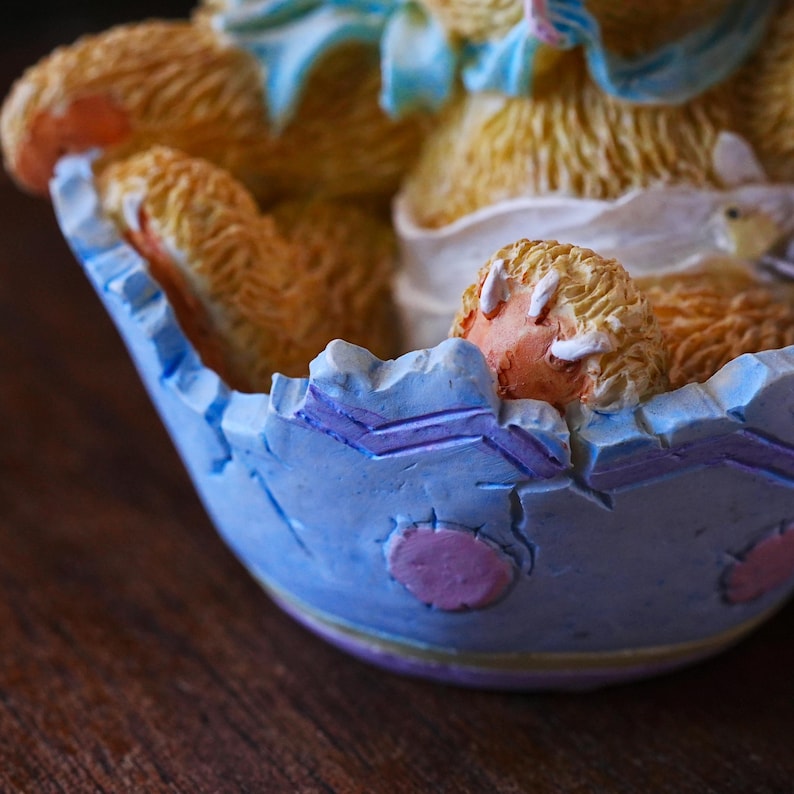 May include: A decorative figurine of a teddy bear emerging from a cracked blue egg. The bear is light brown with a peach-colored paw. The egg is light blue with purple and pink accents. The figurine is on a dark brown surface.