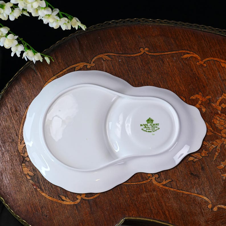 Puede incluir: Plato de porcelana Royal Albert blanco con borde festoneado y un peque&ntilde;o compartimento redondo. El plato tiene un dise&ntilde;o floral y las palabras "Royal Albert Bone China England" est&aacute;n impresas en la parte inferior.