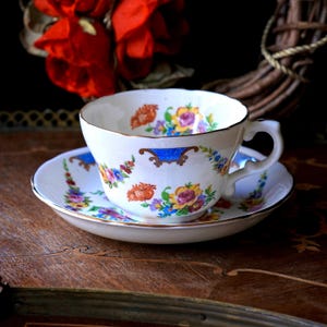 Puede incluir: Un juego de taza y platillo vintage. La taza y el platillo de porcelana blanca están adornados con coloridos diseños florales en naranja, amarillo, morado y azul. La taza tiene un borde dorado y un acento azul.
