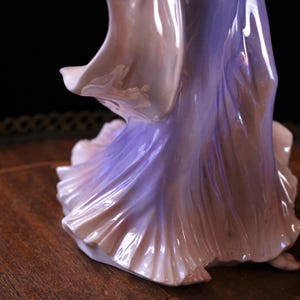 May include: A porcelain figurine of a woman in a flowing gown. The dress transitions from a soft pink to a lavender hue, with iridescent highlights. The figurine's form is detailed, with folds and draping of the fabric.