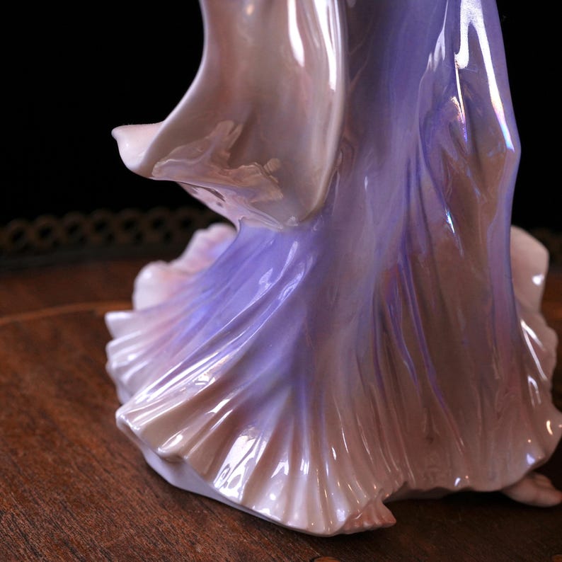 May include: A porcelain figurine of a woman in a flowing gown. The dress transitions from a soft pink to a lavender hue, with iridescent highlights. The figurine's form is detailed, with folds and draping of the fabric.