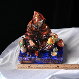 May include: A ceramic figurine of a robed figure with small, colourful pottery. The figure sits on a blue base. The robe is a deep red-brown colour. A ruler shows measurements in centimetres.