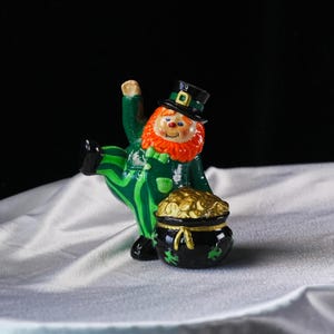 May include: A ceramic leprechaun figurine with a red beard, green suit, and black hat. The figurine is holding a pot of gold coins. The pot is black with green shamrocks. The leprechaun is posed with one leg raised.
