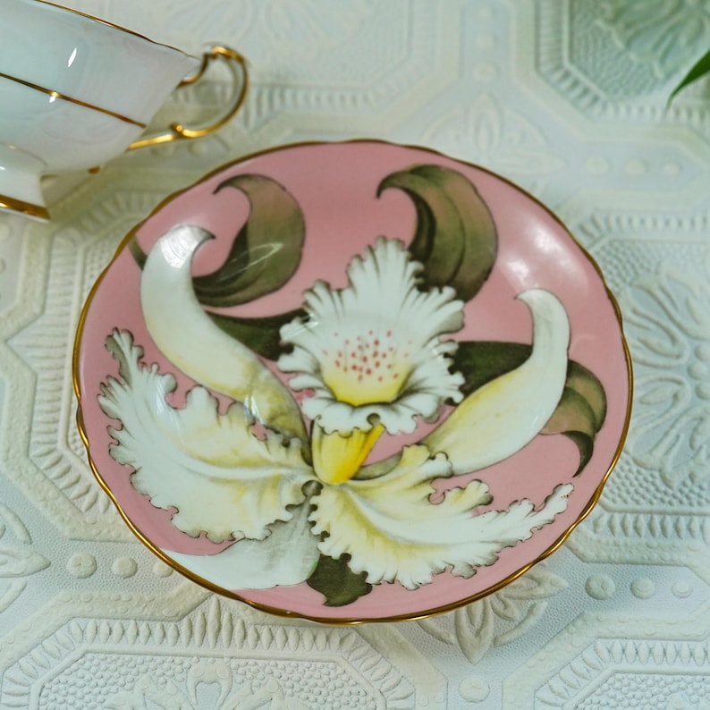 May include: A pink porcelain saucer with a hand-painted orchid design. The orchid features white petals with yellow and green accents, set against a pink background. The saucer has a gold trim and is part of a tea set.