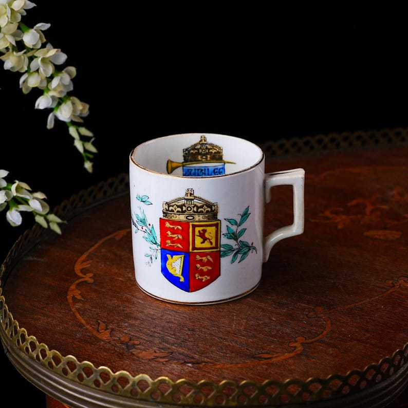 May include: A white ceramic mug with a gold rim and a rectangular handle. The mug features a colorful coat of arms design with a crown and a trumpet. Decorative floral accents are on the sides.