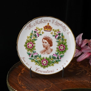 Op de afbeelding: Een wit keramieken herdenkingsbord met een centraal portret van koningin Elizabeth II, een kroon en een bloemenkrans van rozen, distels en klaverbladeren. De tekst luidt H.M. Queen Elizabeth II, Coronation, June 2nd, 1953.