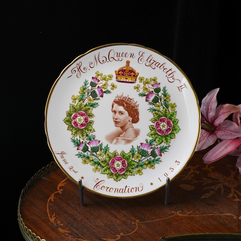 Op de afbeelding: Een wit keramieken herdenkingsbord met een centraal portret van koningin Elizabeth II, een kroon en een bloemenkrans van rozen, distels en klaverbladeren. De tekst luidt H.M. Queen Elizabeth II, Coronation, June 2nd, 1953.