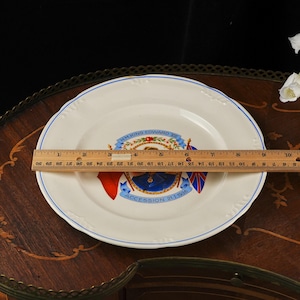 K&ouml;nnte beinhalten: Ein wei&szlig;er Keramikteller zur Erinnerung mit einem Portr&auml;t von K&ouml;nig Edward VIII., blauen B&auml;ndern und Flaggen. Ein &uuml;ber die Mitte gelegtes Holzlineal zeigt einen Durchmesser von etwa 23 cm mit einem blauen Randdetail am Tellerrand.