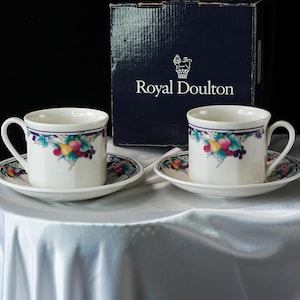 Może przedstawiać: Dwie białe filiżanki i spodki Royal Doulton z owocowym wzorem. Filiżanki i spodki mają niebiesko-fioletową obw&oacute;dkę. Filiżanki i spodki stoją na białym obrusie. Granatowe pudełko z napisem "Royal Doulton" znajduje się w tle.