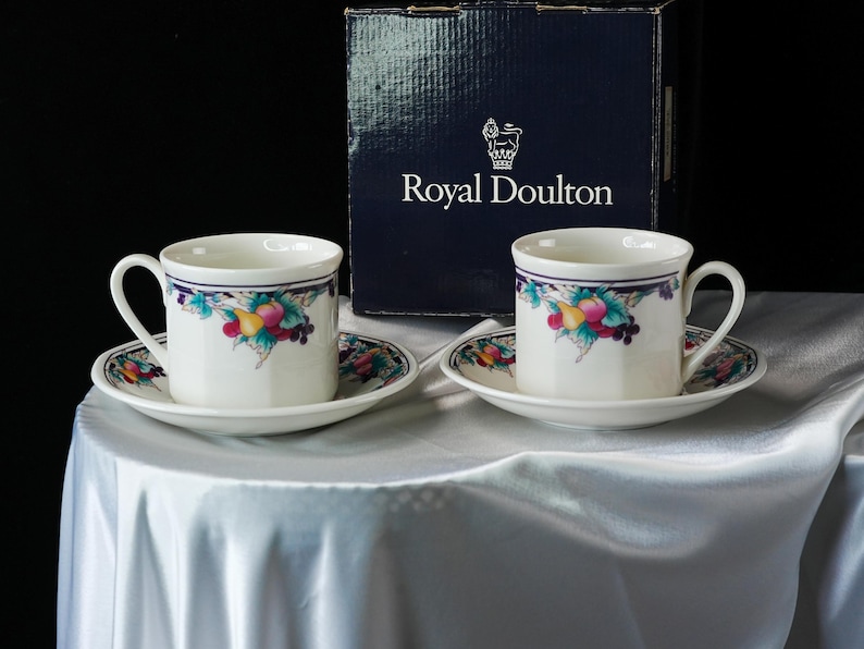 Może przedstawiać: Dwie białe filiżanki i spodki Royal Doulton z owocowym wzorem. Filiżanki i spodki mają niebiesko-fioletową obw&oacute;dkę. Filiżanki i spodki stoją na białym obrusie. Granatowe pudełko z napisem "Royal Doulton" znajduje się w tle.