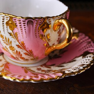 May include: A close-up of a pink and gold teacup and saucer set. The teacup and saucer have a scalloped edge and are decorated with gold floral designs. The handle of the teacup is gold. The teacup and saucer are on a dark surface.