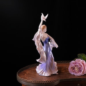 May include: A porcelain figurine of a woman in a flowing lavender and blue gown, holding a white dove. The figurine has a graceful pose with flowing fabric and is set against a dark background. A pink rose is on the right.